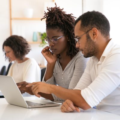 Instructor explaining corporate software specific to intern. Man and woman in casual sitting at desk in classroom, working on laptop, pointing at screen, talking. Education concept